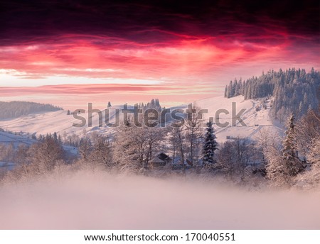Foggy winter landscape in mountain village under the dark red sky
