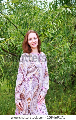 Portrait of a beautiful European woman in a lilac dress with patterns on a background of green trees.
