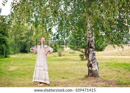 Beautiful young woman in the Russian national dress dancing in the meadow next to the birch tree.