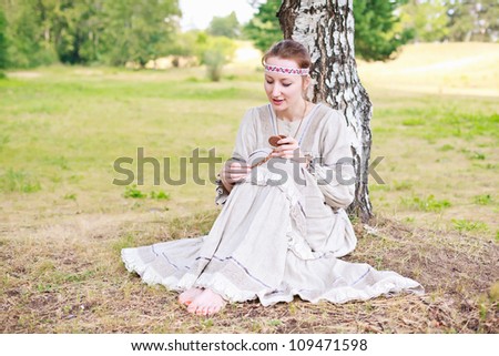 Young beautiful woman in the Russian national dress sitting on a meadow next to the birch tree.