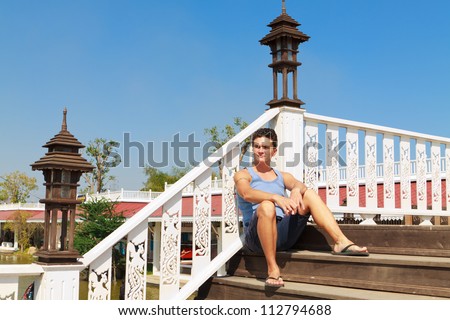 A young white man with short hair sitting on the stairs of the wooden bridge with white railing against the red roofs at the floating market in Thailand.