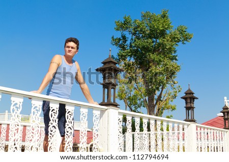 A young white man with short hair, standing on a wooden bridge with white railings against the clear blue sky.