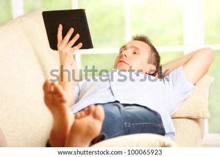 Man relaxing with tablet, laying on sofa in home with big window in background