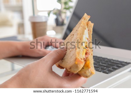 Woman eating a breakfast sandwich and drinking coffee while working with a laptop