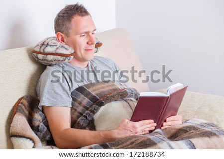 Man sitting comfortably with neck pillow and woolen blanket, reading book.
