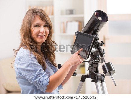 Beautiful woman with astronomical telescope standing near a window and checking something about stars and planets on the tablet