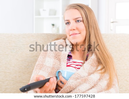 Bored young woman covered with blanket holding cup and remote control and watching tv at home