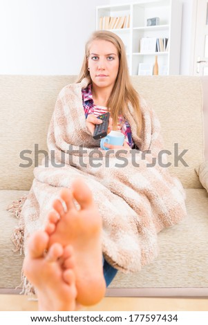Bored young woman covered with blanket holding cup and remote control and watching tv at home