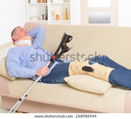 Man with leg in neck brace, knee cages and crutches for stabilization and support resting on a sofa