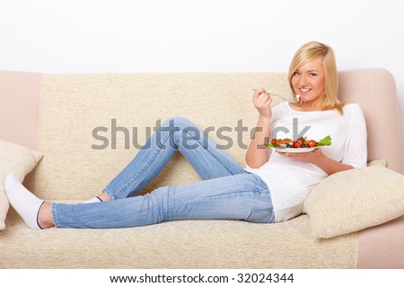 Beautiful woman sitting on the sofa and eating a healthy Greek salad of tomatoes, mozarella cheese, olives, lettuce