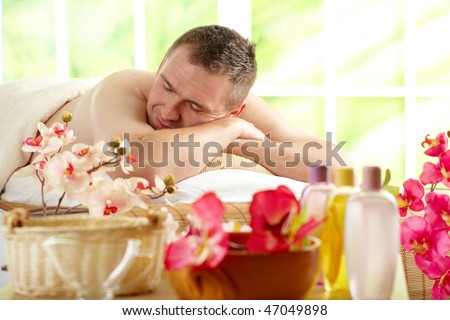 Man relaxing in sunny spa salon with closed eyes laying on the mat with flowers and cosmetics