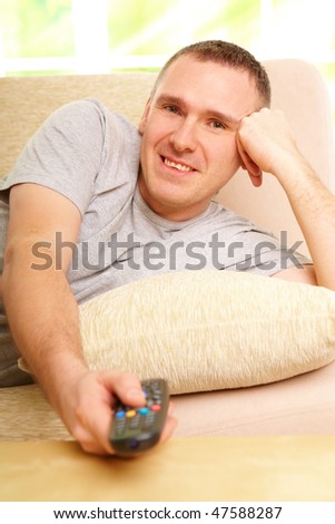 Relaxed handsome man smiling lying on sofa with remote control handheld, watching television in home