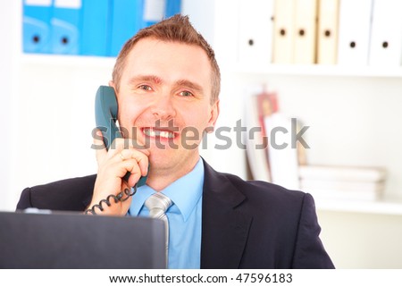 Happy businessman sitting in office, talking on land-line phone and smiling. Documents in background.