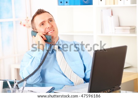 Happy businessman sitting at desk in office, talking on land-line phone with laptop and smiling. Documents in background.