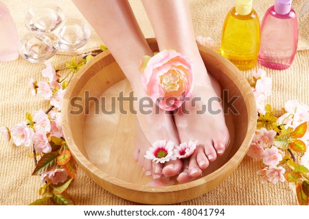 Close-up of beautiful female feet in wooden bowl filled with fluid cosmetics, flowers all around. Concept of natural spa treatment.