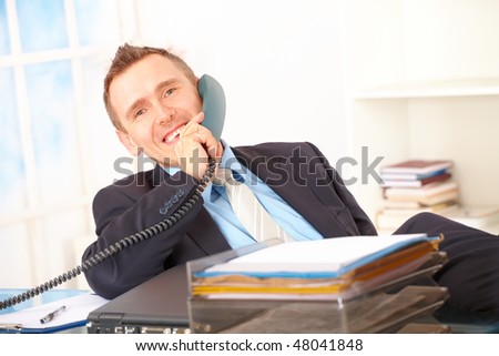 Happy businessman sitting in office, talking on landline phone and smiling with documents and laptop on his desk