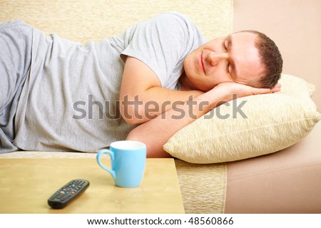 Portrait of a man sleeping on sofa in home with pillow under head. Blue cup and TV controller in foreground.