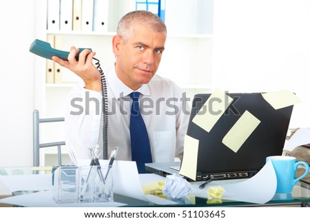 Stressed overworked mature businessman sitting at desk with phone and laptop with many note stickers