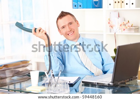 Happy businessman sitting at desk in office, talking on landline phone with laptop and smiling. Documents in background.