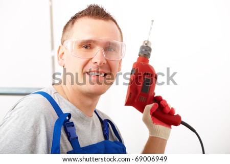 Cheerful man working with red drill, wearing protective glasses, blue trousers and gloves, ladder in background