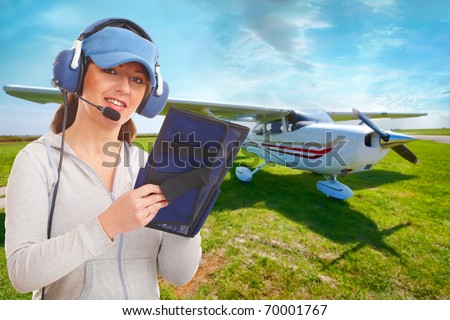 Cheerful woman pilot with headset used in aircraft taking notes on knee-pad, with airplane in the background