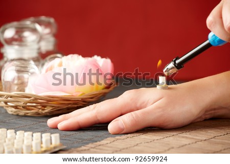 TCM Traditional Chinese Medicine. Hand lighting mini moxa stick, flower and natural herbs in glass jars in background.