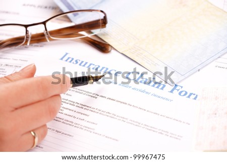 Hand filling in insurance claim form. Other papers like ID or vehicle documents and glases in the background