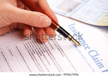 Hand filling in insurance claim form. Other papers like ID or vehicle documents in the background
