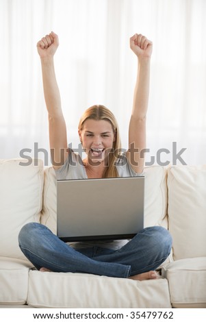 An excited looking young blonde raising her fists in the air and cheering.  She is smiling and has a laptop computer in her lap.  Vertically framed shot.