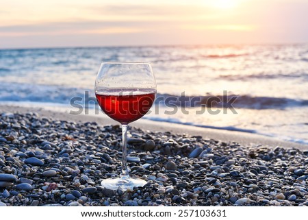 Romantic glass of wine sitting on the beach at colorful sunset, Glass of red wine against sunset, red wine on the sea ocean beach, sky background with clouds