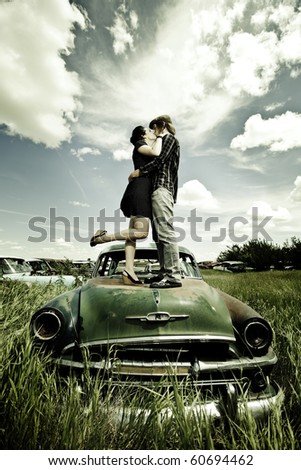 A young couple are kissing on a old broken car in a field./Young Couple Kissing on Old Broken Car