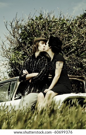 A young couple are kissing on a old broken car in a field. /Young Couple