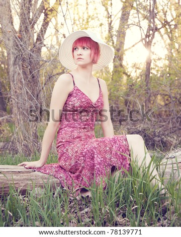 An attractive red haired woman sitting on a log wearing a sundress.