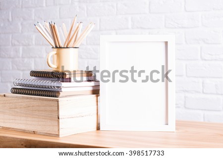 Desk with blank white picture frame and pencils in iron mug placed on books and wooden box. Mock up