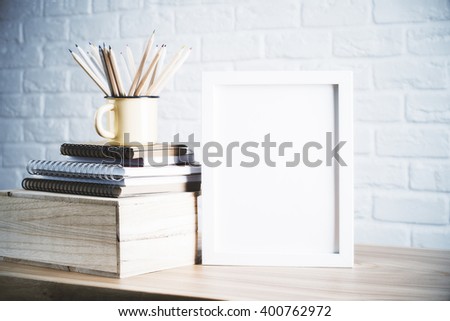 Desk with blank picture frame and pencils in iron mug placed on books and wooden box. Mock up