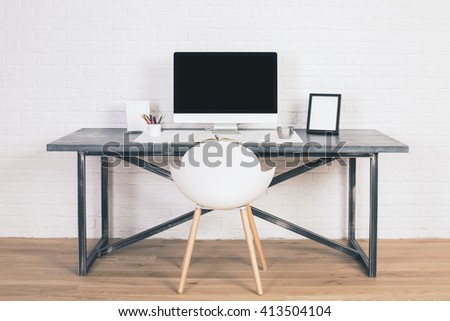 Front view of designer desk with blank computer monitor, frames and other items with white chair next to it. Wooden floor and white brick wall background. Mock up