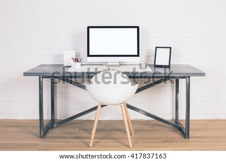 Front view of designer desk with blank white computer screen, frames and other items with white chair next to it. Wooden floor and white brick wall background. Mock up
