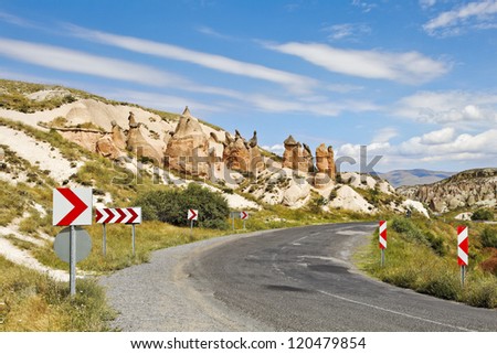 Landscape of Cappadocia rural country lane lined with natural geological volcanic activity, rock formation, limestone, sandstone, chevrons, bend in the road, striking blue sky and clouds