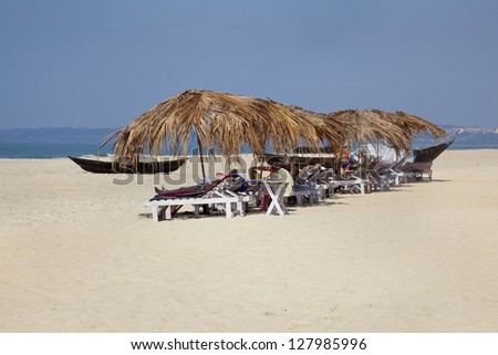 Landscape of secluded beach and a row of tropical parasols with day trippers and holidaymakers basking in the sun under the shade. White sand, blue sky, sea and fishing boats for backdrop in Goa India