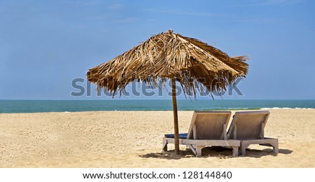 Panorama of a generic tropical deserted beach with loungers under a coconut leaf parasol. Blue sky, ocean, gentle surf and clean sandy beach. Location, Goa India
