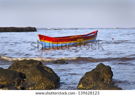 Horizontal early morning landscape of brightly colored fisherman canoe moored on the coastline of the Arabian Sea at Manori, Bombay, India with surf and waves lapping on nearby rocks