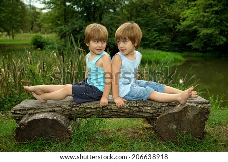 Adorable Little Twin Brothers Sitting on a Wooden Bench, Smiling and Looking at Each Other Near the Lake at Summer