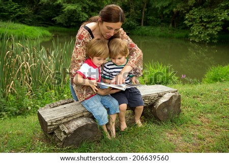 Charming Mother Reading a Book to Her Adorable Little Twin Sons While Sitting Outside  Near Beautiful Lake at Summer