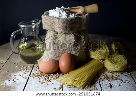 Pasta, Two Eggs, Jute Bag Filled with Flour, Wooden Spoon and Olive Oil in Glass Bottle on an Old Wooden Table, Black Background