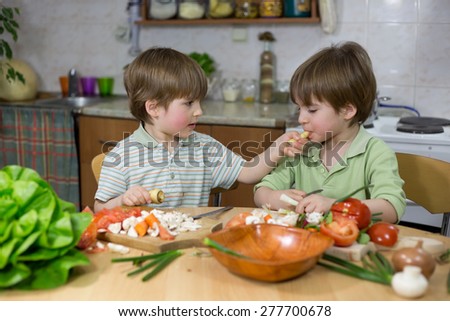 Cute Little Boy Feeding His Twin Brother With Parsnip at Kitchen Table at Home