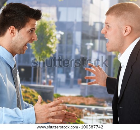 Angry businessmen standing facing each other, shouting and gesturing in front of office.