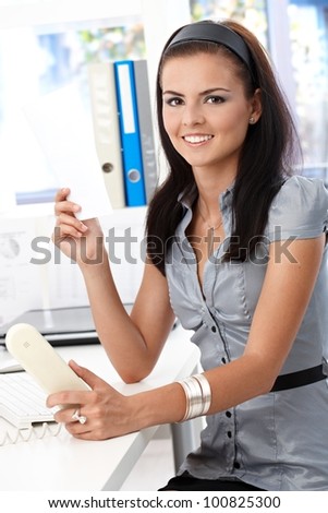 Young attractive secretary working in bright office, smiling, looking at camera.