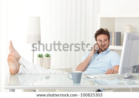 Cool man with feet up on desk making mobile phone call, typing on keyboard, smiling, looking at computer screen.