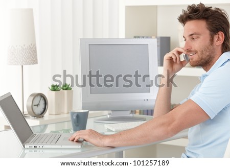 Smiling man talking on mobile phone while using laptop computer at desk in study. Blank space on screens for your logo or image.