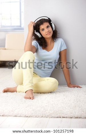 Attractive young woman listening music through headphones, sitting on floor, smiling.